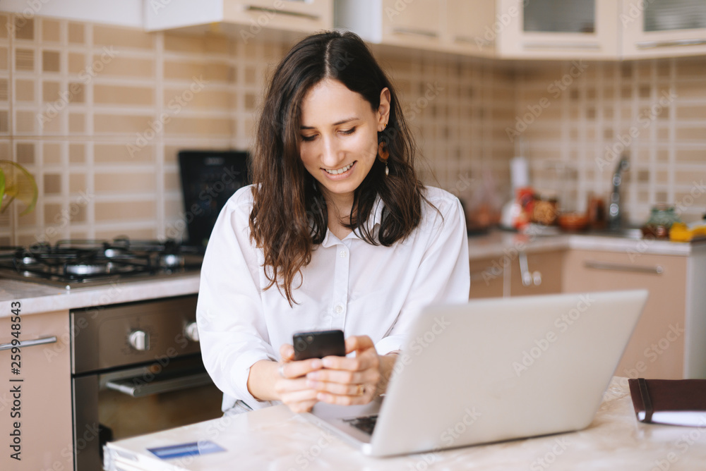 Beautiful smiling woman using phone while working on laptop in kitchen room Beautiful smiling woman using phone while working on laptop in kitchen room