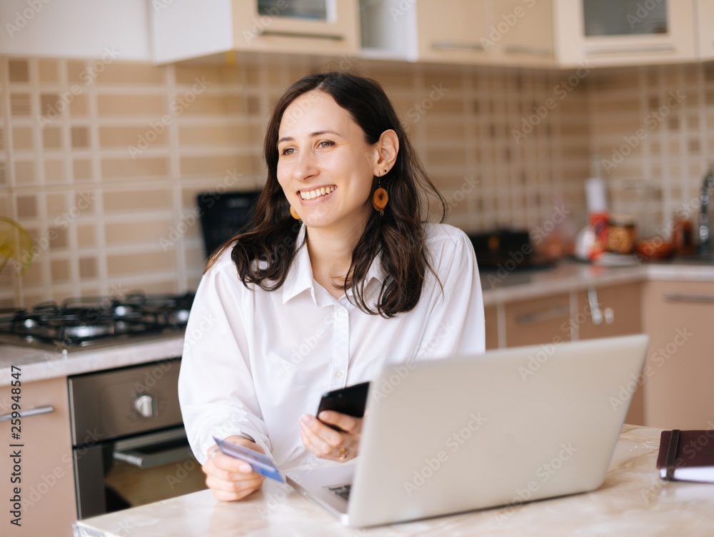 Photo of happy young woman with laptop, credit card and smartphone looking forward in kitchen at home