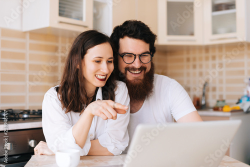 Photo of young happy couple sitting in kitchen and using laptop together, Family concept