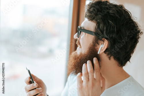 Handsome bearded businessman is using mobile and airpods, while talking with coworker near window photo
