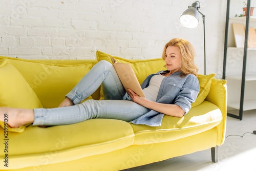 Woman lying on cozy yellow couch and reading book