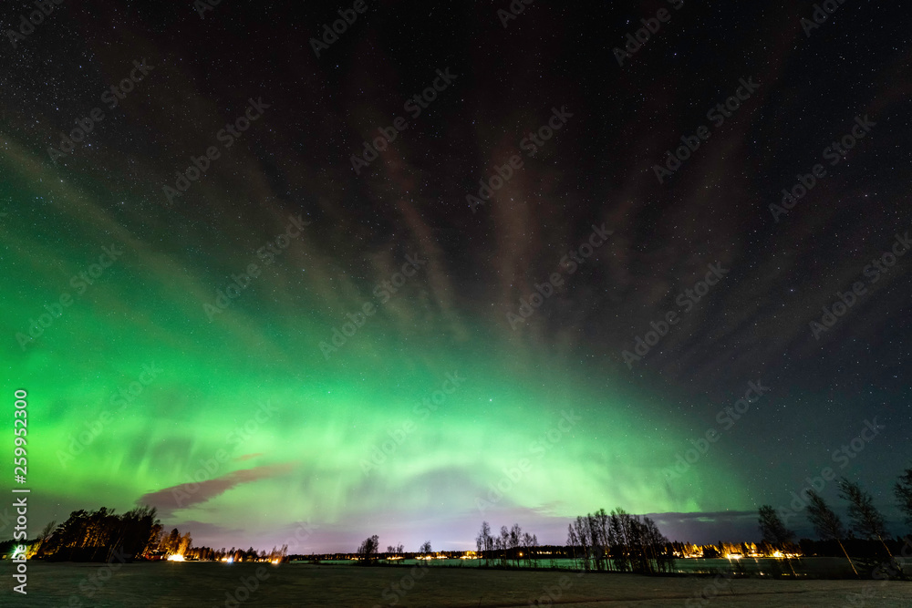 Aurora at clear skies with very tiny clouds shines above a field and lake at Scandinavian countryside, winter time