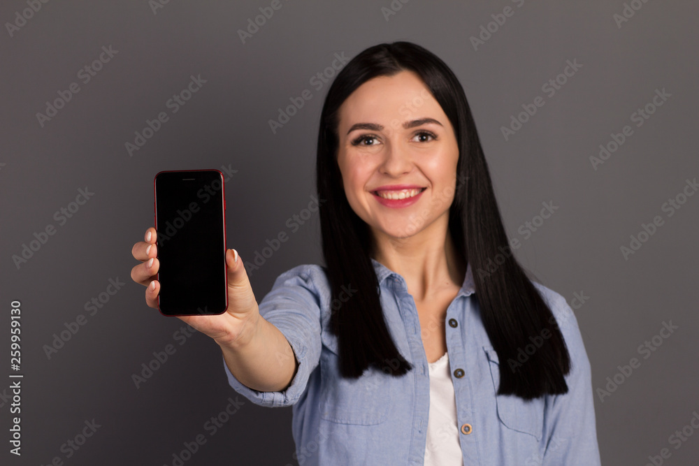 Closeup photo studio portrait of a pretty cute with a toothy smile holding a phone in hand device gadget wearing denim shirt isolated gray background copyspace Closeup photo studio portrait of a pretty cute with a toothy smile holding a phone in hand device gadget wearing denim shirt isolated gray background copyspace