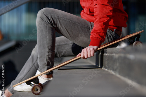 Stylish man longboarder in casual clothes resting on the steps, sitting with longboard/skateboard outdoors, side view, cropped image. Urban, subculture, skateboarding concept