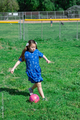 Little Girl in Blue Dress kicking soccer ball on Green Grass