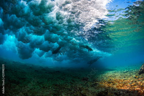 Powerful ocean wave breaks over the coral reef. There are underwater photographer and female surfer diving under the wave in the frame
