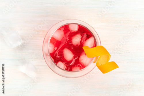 A cocktail with campari and an orange curl garnish, shot from above on a white wooden background with ice cubes and a place for text photo