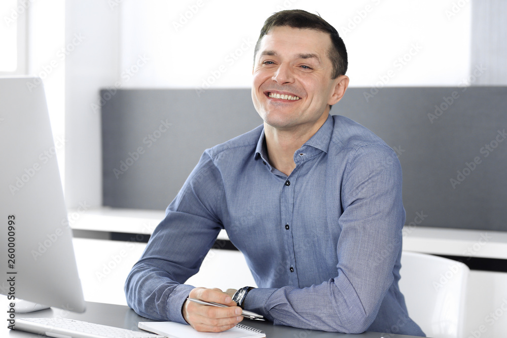 Cheerful smiling businessman working with computer in modern office. Headshot of male entrepreneur or director of a company at the workplace. Business concept 