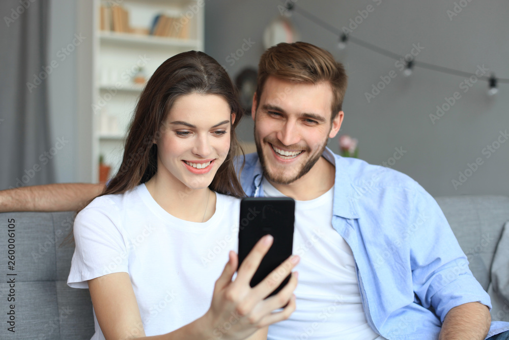Young couple watching online content in a smart phone sitting on a sofa at home in the living room. Young couple watching online content in a smart phone sitting on a sofa at home in the living room.
