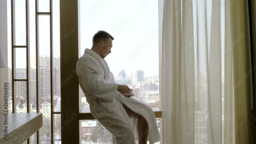 Handsome man dressed in white bathrobe drinking coffee and looking out of the window at home. 4K Handsome man dressed in white bathrobe drinking coffee and looking out of the window at home. 4K
