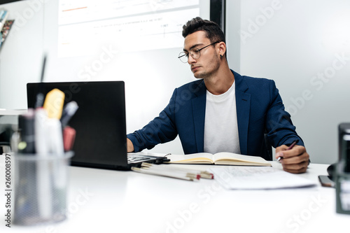 Dark-haired young architect in glasses and in a blue jacket is working with documents on the desk in the office