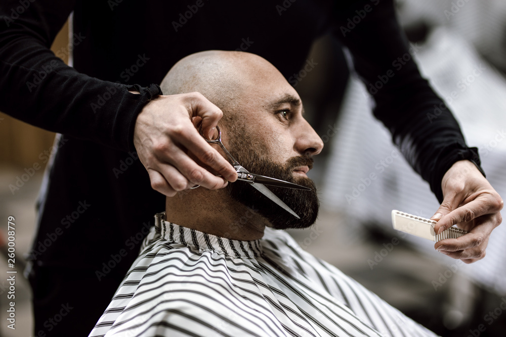 Fashion barber dressed in a black clothes scissors beard of brutal man in the stylish barbershop