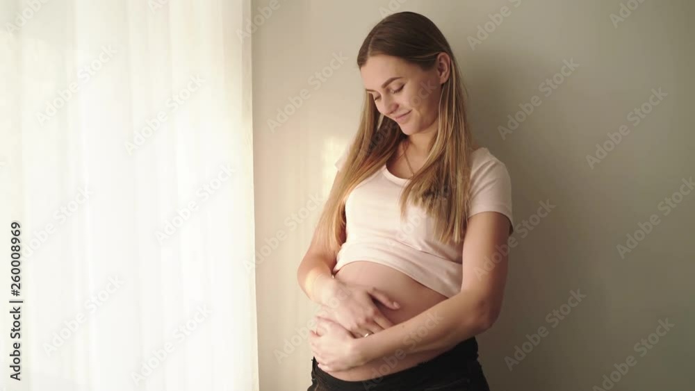 Happy pregnant woman caresses her belly at window in room Happy pregnant woman caresses her belly at window in room