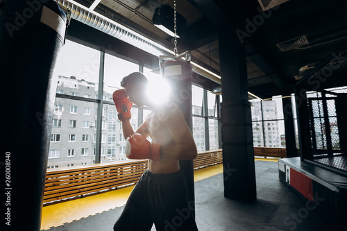 Sportsman in red boxing gloves with a naked torso dressed in the black shorts hits punching bag in the gym with panoramic windows