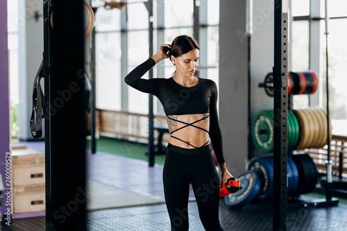 Athletic dark-haired girl dressed in black sportswear stands with water in her hand near the sport equipment in the gym and touches hair