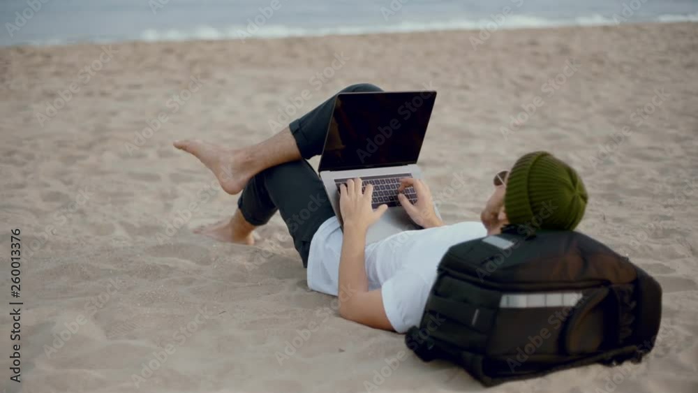 Young millennial freelancer or office worker relaxes chill on beach with laptop on lap, works remotely from work and home during vacation or holiday, workaholic addiction