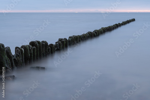 Long exposure photo of a wooden wavebreaker in the evening. Port or ship lights visible in the background.