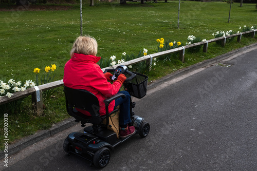 An elderly lady in a red coat enjoying the freedom of an electric mobility scooter on a quiet road with spring daffodils in bloom.