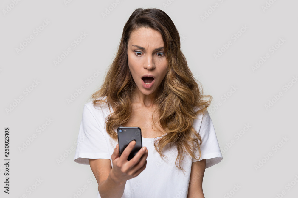 Amazed shocked woman over grey background looking on smartphone screen Amazed shocked woman over grey background looking on smartphone screen