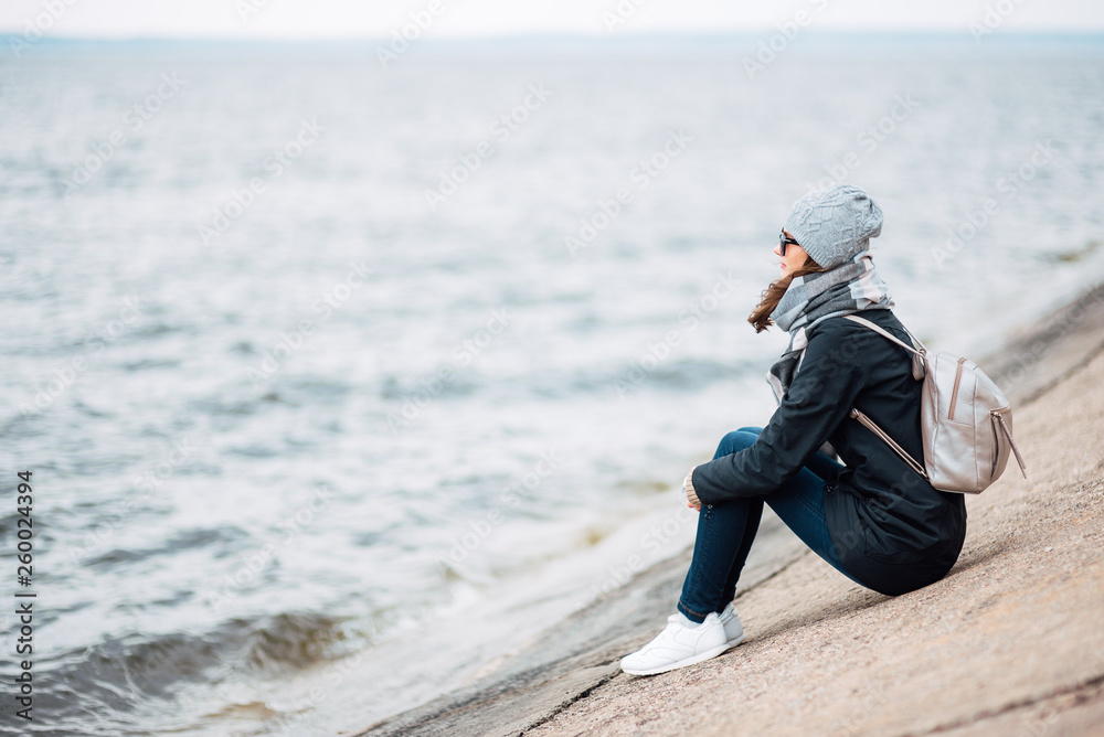 young woman traveler sits near the beach in the fall in windy weather. full length photo of girl in coat, hat, backpack and large scarf outdoors young woman traveler sits near the beach in the fall in windy weather. full length photo of girl in coat, hat, backpack and large scarf outdoors