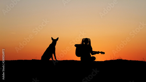 Silhouette girl guitarist on sunset background, silhouette of a dog breed Belgian Shepherd Malinois, happy friends, outdoor © Diana Badmaeva