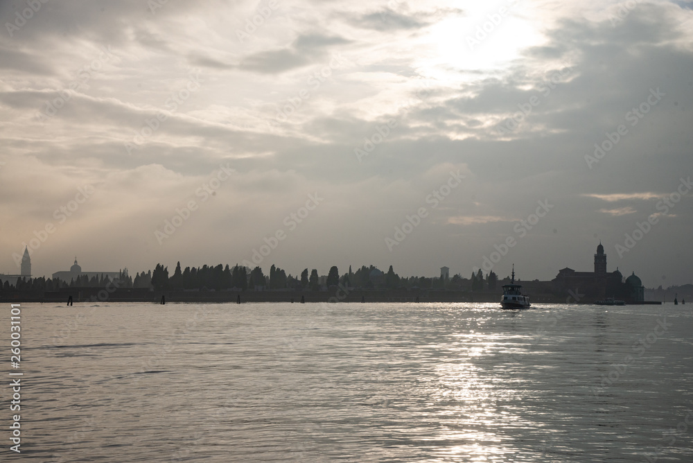 Venice lagoon with black clouds in the sky. You see a ferry, the cemetery island with the church of San Michele in Isola also known as San Michele di Murano and the profile of Venice. Cold rain-laden 