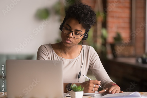 Focused mixed race woman wearing headphones watching webinar write notes photo