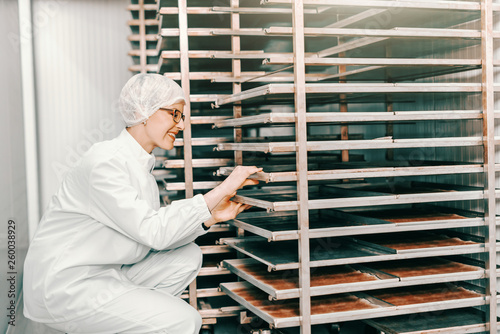Young female Caucasian blonde employee in sterile uniform putting trays on rack while crouching in food factory.