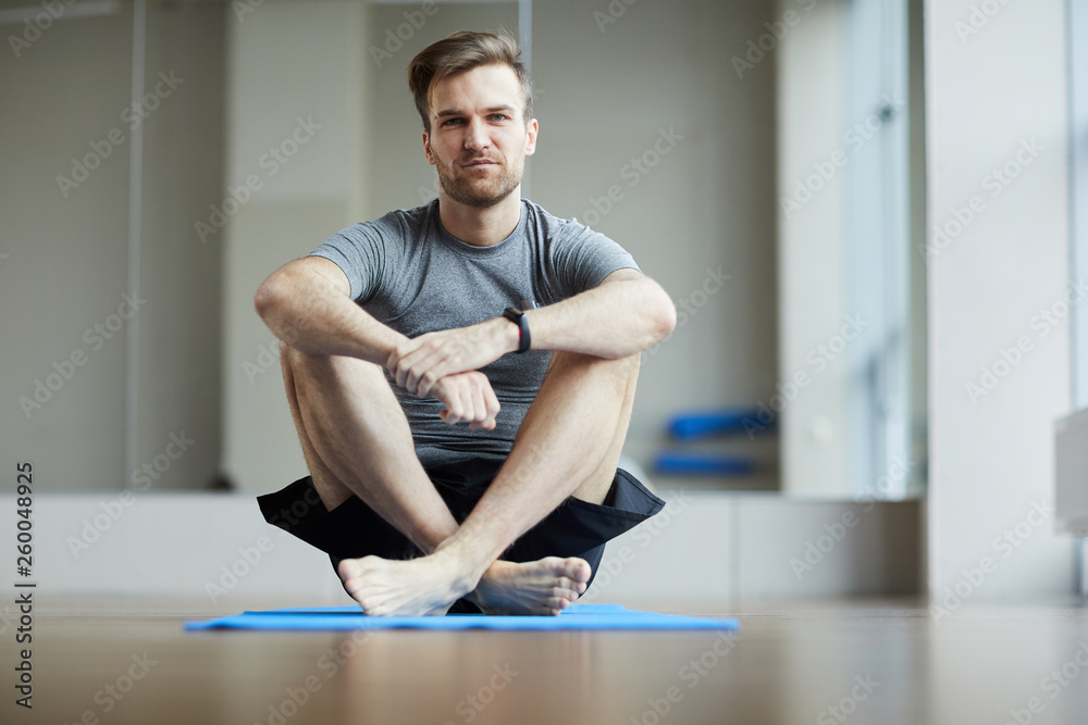 Fototapeta premium Serious pensive young man sitting on exercise mat and embracing knees while waiting for yoga class, he looking at camera