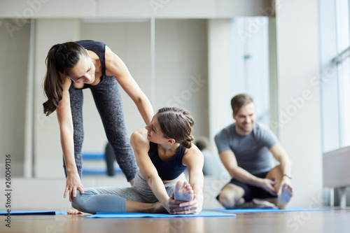 Cheerful kind young yoga teacher assisting girl and pushing her back toward to make stretching exercise more effective