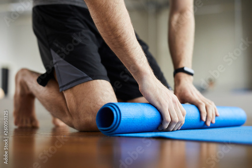 Close-up of unrecognizable man standing on knees and rolling out exercise mat after yoga training in gym 