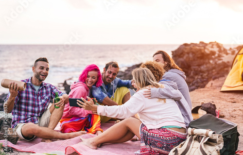 Happy friends doing party at sunset while camping next the beach - Millennial young people having fun drinking beer and using mobile smartphone outdoor - Summer, vacation, tech and youth concept
