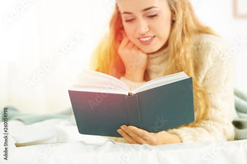 Attractive young woman in cozy warm sweater reading book on bed at home. Space for text