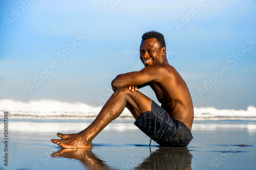 young attractive and happy black afro American man with fit muscular body sitting on beach sand enjoying beautiful view smiling cheerful in Summer holidays