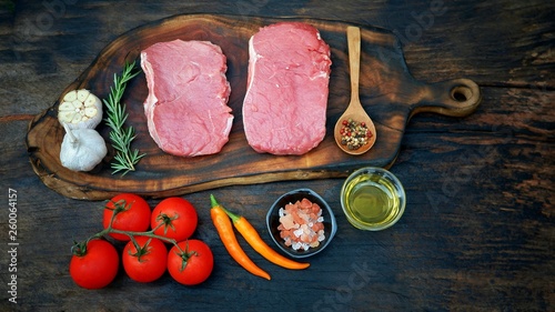 Preparation for steak cooking, fresh raw meat beef steak, rosemary,pepper,garlic,cherry tomatoes,chilli,salt and vegetable oil lined on wood cutting board and dark table floor background