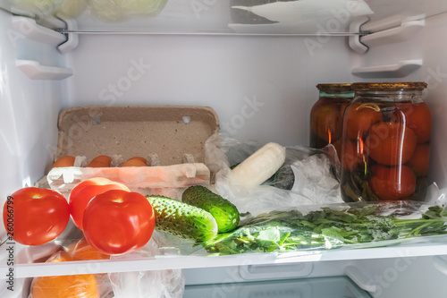 Shelf in the refrigerator, filled with cans of canned tomatoes, as well as vegetables, herbs, cheese. The image is suitable for themes of food choices, diet, home cooking.