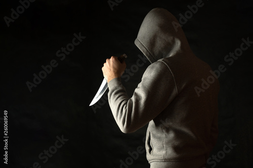 A man in a hood on a black background, brandishing with a knife in his hand. A criminal with a knife attacks from the dark.