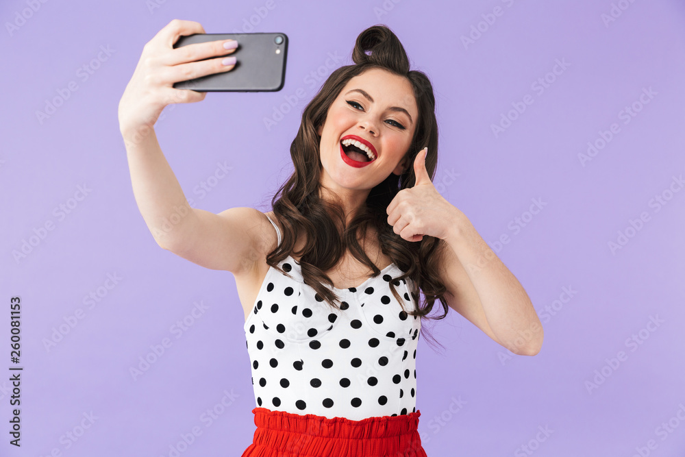 Naklejka premium Photo of beautiful pin-up woman 20s in vintage polka dot dress holding and taking selfie photo on black smartphone