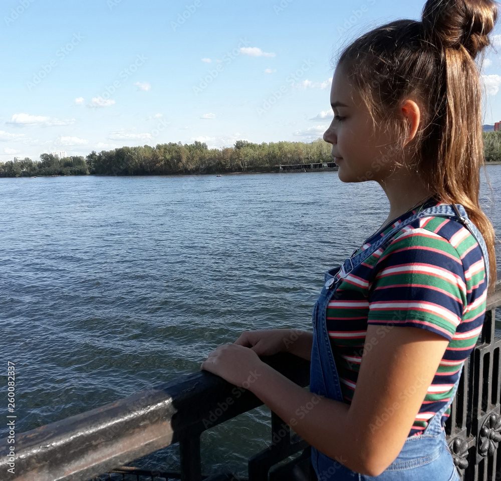 A young girl on the embankment of a large river looks at the water. Long hair gathered in a bun. Summer, striped short-sleeved clothing, jeans.