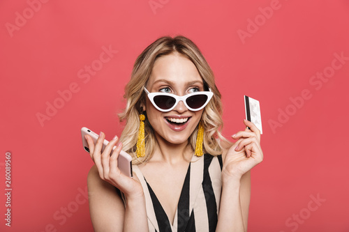Beautiful amazing young woman posing isolated over red coral background using mobile phone holding credit card.