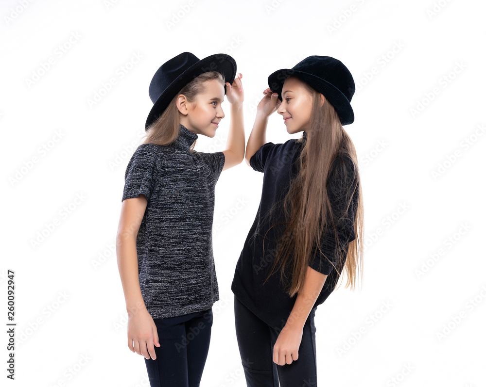 Two cute teenage girlfriends schoolgirls wearing turtleneck sweaters, jeans and hats, smiling dancing and look on each other. Isolated on white. Fashion and advertising design. Two cute teenage girlfriends schoolgirls wearing turtleneck sweaters, jeans and hats, smiling dancing and look on each other. Isolated on white. Fashion and advertising design.