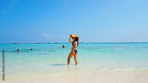 The girl in the hat and swimsuit on the beach. Glasses on. European woman on the beach with white sand. The water is turquoise with a blue tint. Photo shoot model. White sand and blue sky. Paradise.