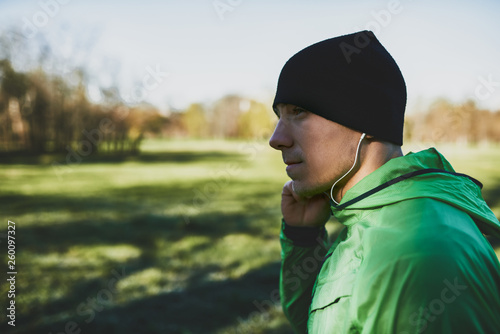 Side view of close up portrait of healthy sportive young man standing outside in park after jogging, listening the music on earphones. Confident young man ready of workout.