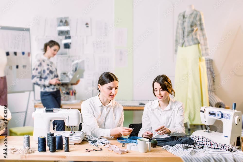 Fashion Design, dressmaker, tailor and fashion concept. Two young women dressmakers sewing while sitting at their working place. Fashion designer on the background creating new fashionable styles. Fashion Design, dressmaker, tailor and fashion concept. Two young women dressmakers sewing while sitting at their working place. Fashion designer on the background creating new fashionable styles.