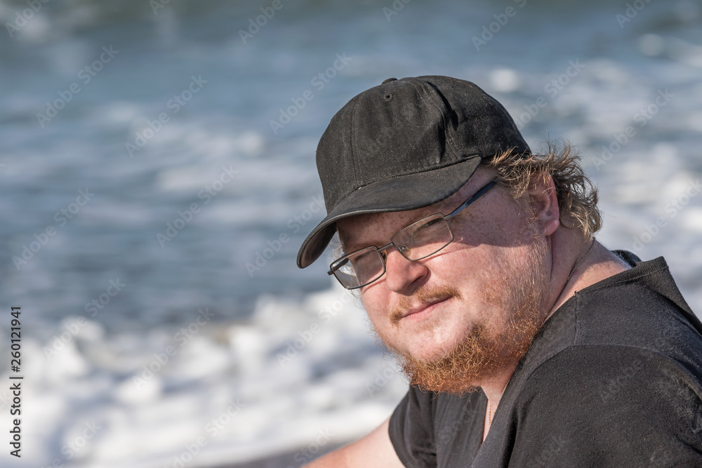 Portrait of a young man with a raw rusty beard dressed in a black shirt and a black cap sitting on the sea shore looking at the camera. Portrait of a young man with a raw rusty beard dressed in a black shirt and a black cap sitting on the sea shore looking at the camera.