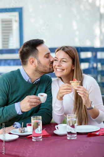 Young couple sitting in a cafe and enjoying coffee time. Man is kissing his girlfriend on cheek, laughing and joking