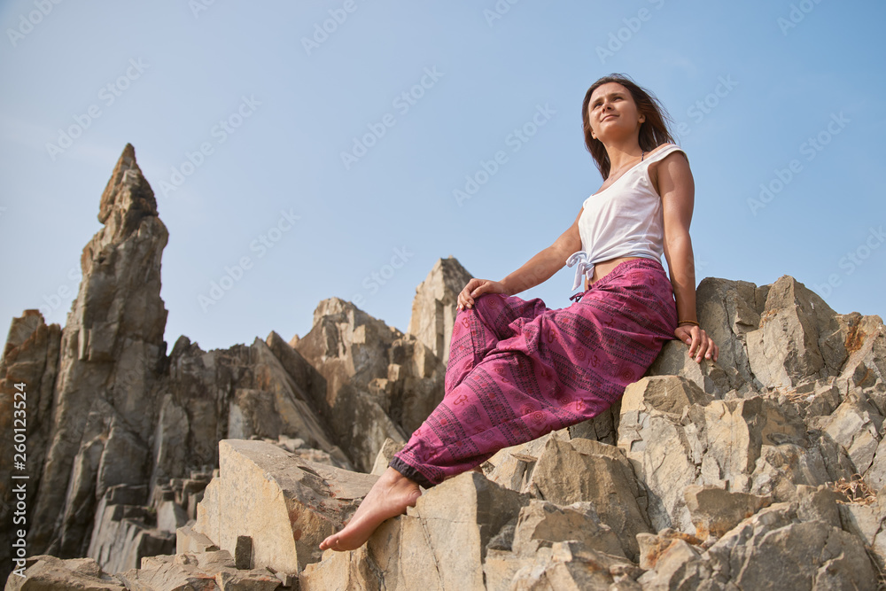 Beautiful young woman with dark hair in vest and wide pants sits on a rock against the sky. The concept of rest in the warm season