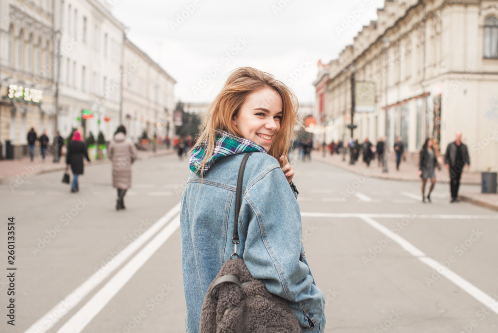 Fototapeta premium Portrait of a smiling girl on the street wearing casual clothes and a backpack, looking in camera and laughing. Stylish lady in a denim jacket smiles and poses on the camera, background of the street
