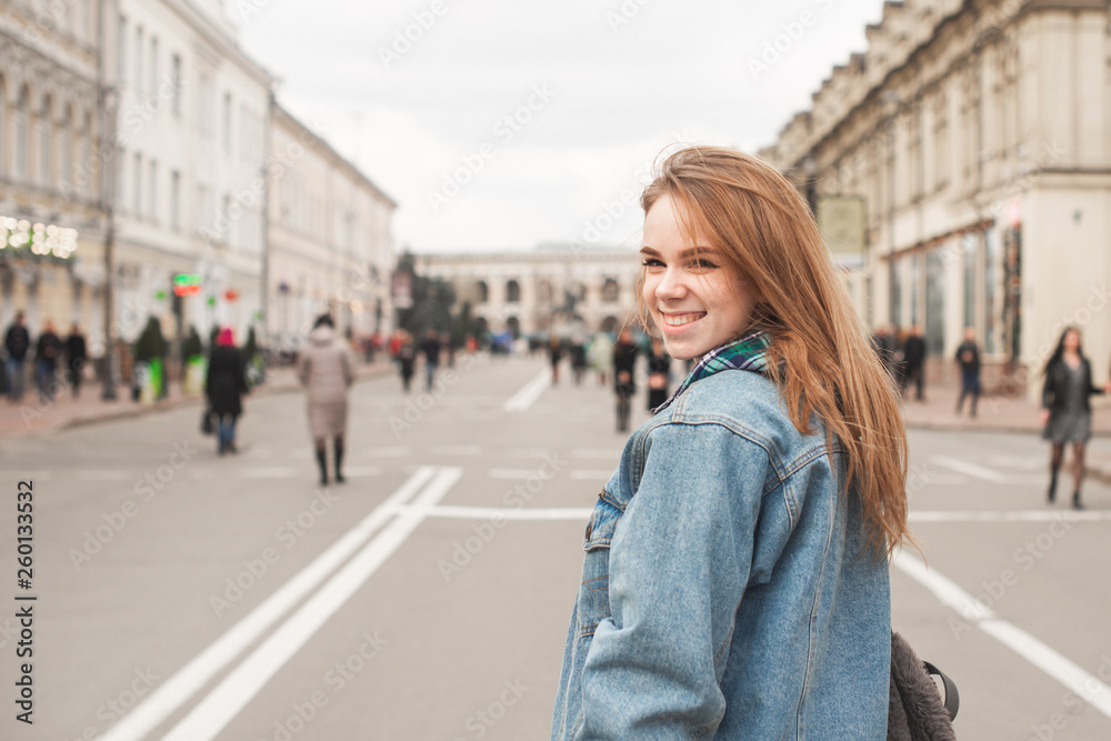 Joyful student girl walking down the street, wearing casual clothes, smiles and looks at the camera. Street girl portrait in denim clothing.