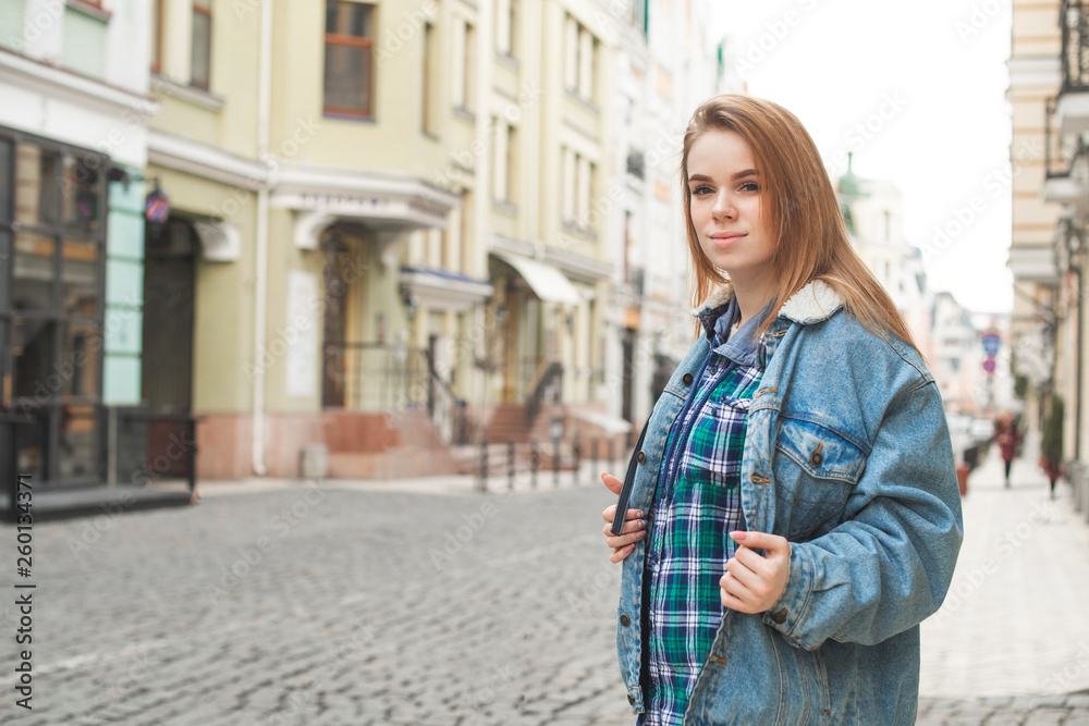 Beautiful lady in casual clothing is standing with a backpack in the background of a street landscape, looking into the camera and smiling. Street portrait of a beautiful stylish girl. Street fashion